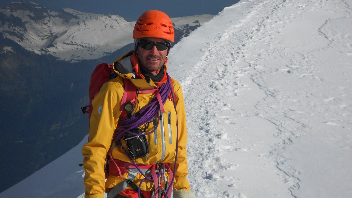 Alpiniste casqué souriant sur une arête enneigée, corde et matériel au baudrier