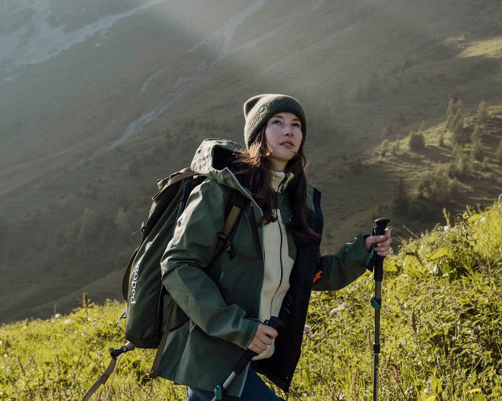 Femme en randonnée avec bâtons dans une prairie de montagne, portant le bonnet en laine Lagoped GEBO CUFF gris zinc, lumière de fin de journée