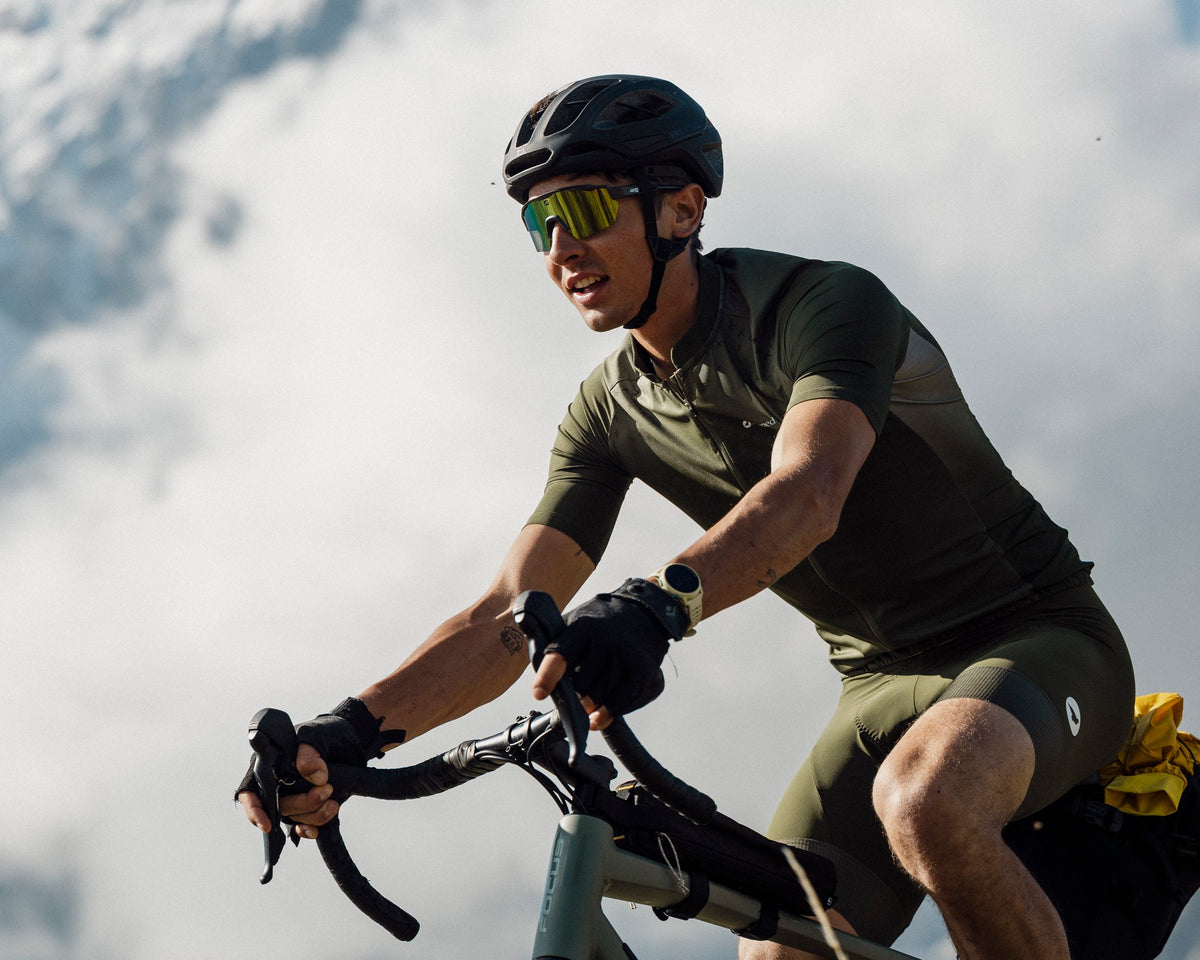 Homme à vélo gravel pédalant en montée devant les nuages, portant le maillot de vélo Lagoped CARDUELIS thym en montagne