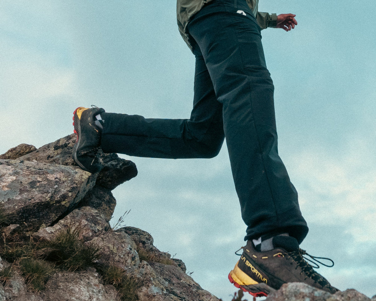 Homme en randonnée portant le pantalon de randonnée Lagoped PTARMIGAN2 black, saut dynamique d'un rocher à l'autre en montagne